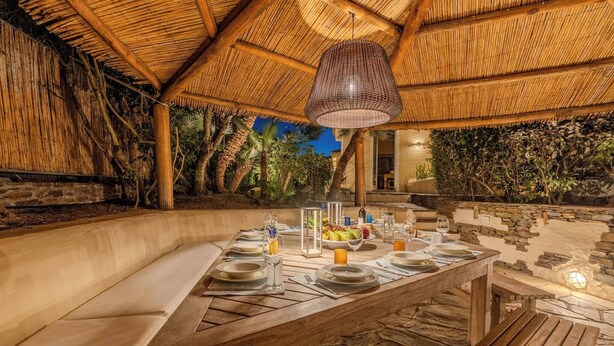 An outdoor dining area with a thatched roof, stone walls, and a wooden table set for a meal. - Villa Flor, Exclusive Property With Pool in Stintino (Stintino)