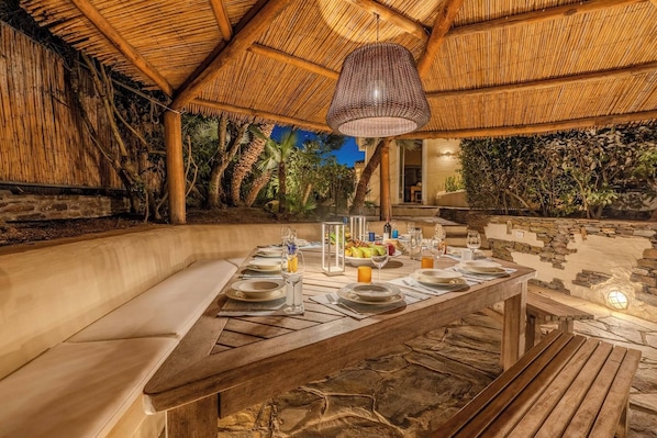 An outdoor dining area with a thatched roof, stone walls, and a wooden table set for a meal. - Villa Flor, Exclusive Property With Pool in Stintino (Stintino)