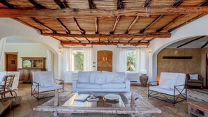 A spacious living room with a high wooden ceiling, a white sofa, two armchairs, a coffee table, and a view of the outdoors through large windows. - Villa Flor, Exclusive Property With Pool in Stintino (Stintino)