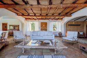 A spacious living room with a high wooden ceiling, a white sofa, two armchairs, a coffee table, and a view of the outdoors through large windows. - Villa Flor, Exclusive Property With Pool in Stintino (Stintino)