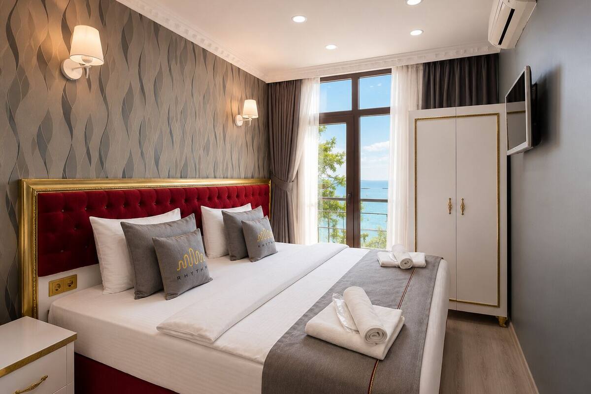 Un dormitorio con cabecera tapizada en rojo, una cama grande con ropa de cama blanca y gris, dos lámparas de pared, un armario y vistas al mar a través de una ventana.