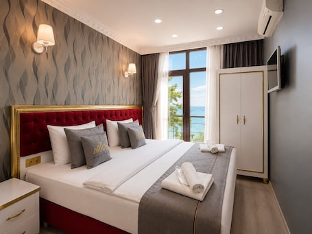 A bedroom with a red tufted headboard, a large bed with white and gray bedding, two wall-mounted light fixtures, a wardrobe, and a view of the sea through a window.