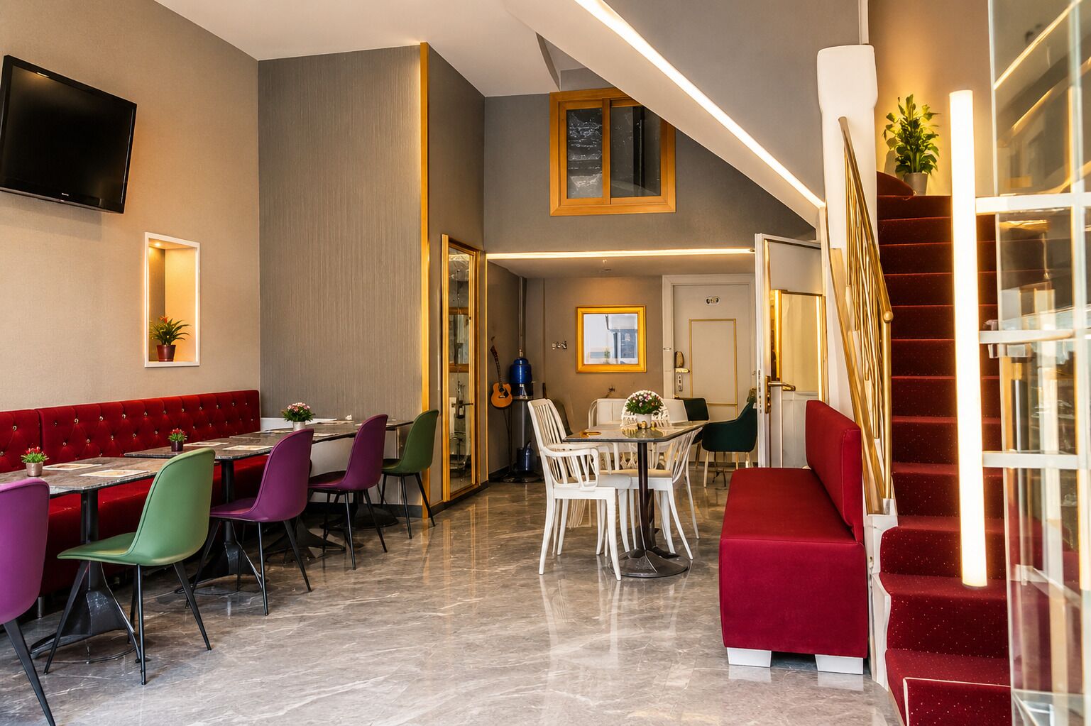 A hotel lobby with a red cushioned seating area, a dining area with colorful chairs, a staircase with red steps, and a flat-screen TV mounted on the wall.
