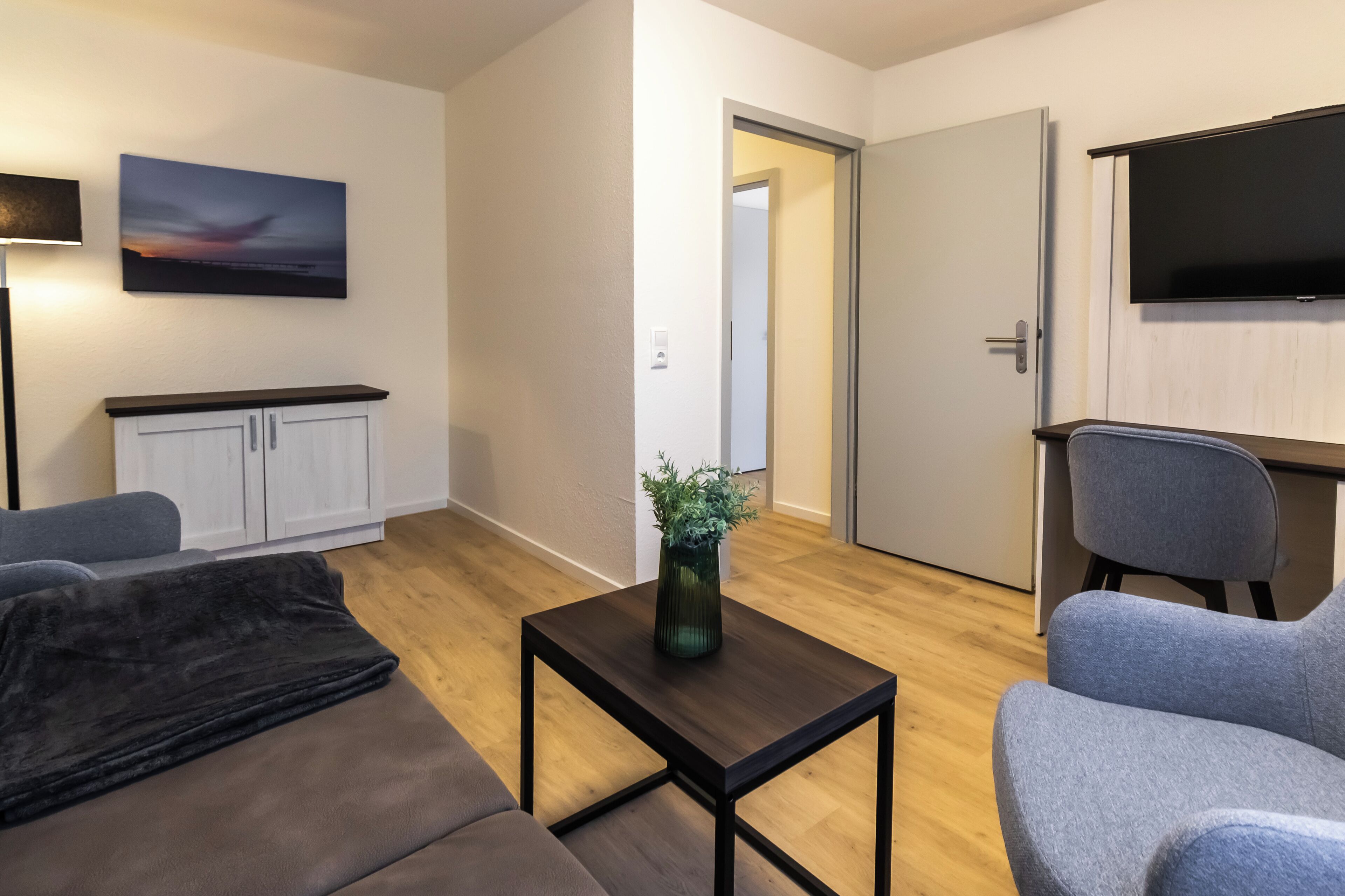 A modern living room with a sofa, a coffee table with a plant, a TV, and a door leading to another room.