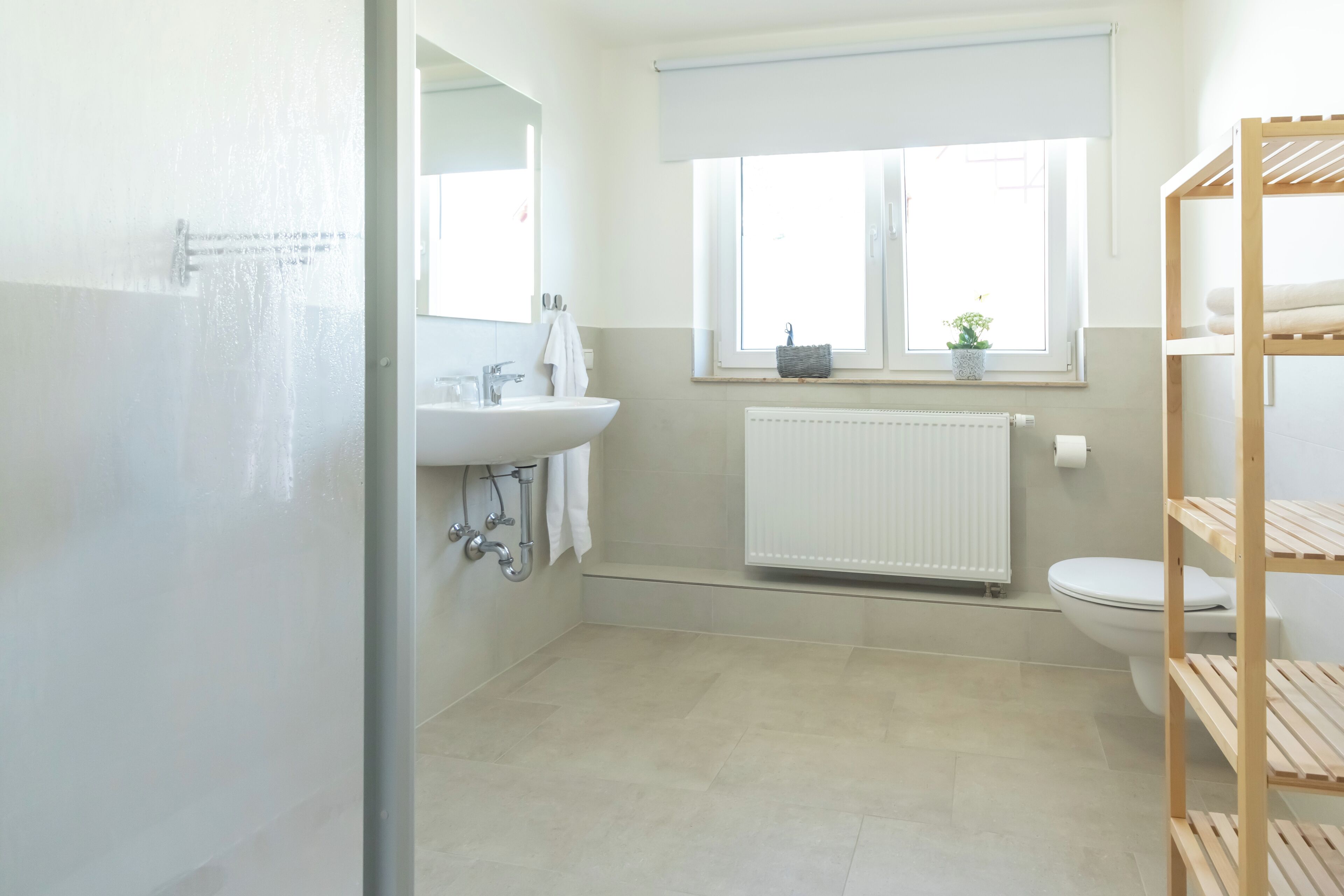 Ein Badezimmer mit weißem Waschbecken, Spiegel, Fenster, Heizkörper und einem Holzregal.