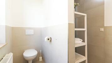 Un baño con inodoro, un rollo de papel higiénico, zona de ducha y un estante con toallas.