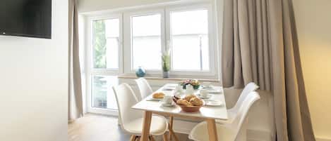 Un coin repas avec une table dressée pour quatre personnes, des chaises blanches, un téléviseur à écran plat fixé au mur et une fenêtre avec des rideaux.