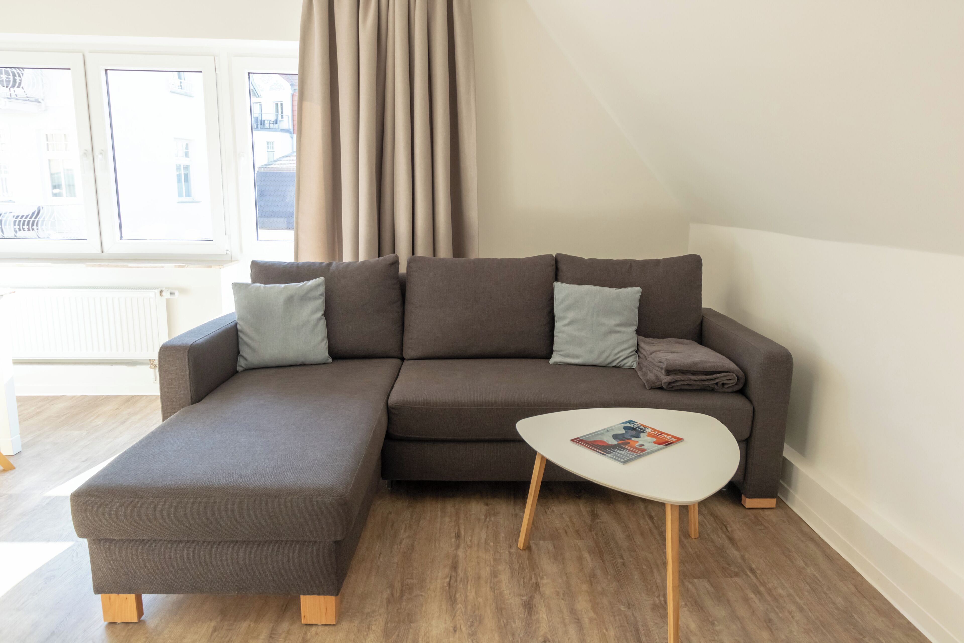 Un salon moderne avec un canapé d’angle, une petite table basse blanche et une vue sur la ville grâce à de grandes fenêtres.