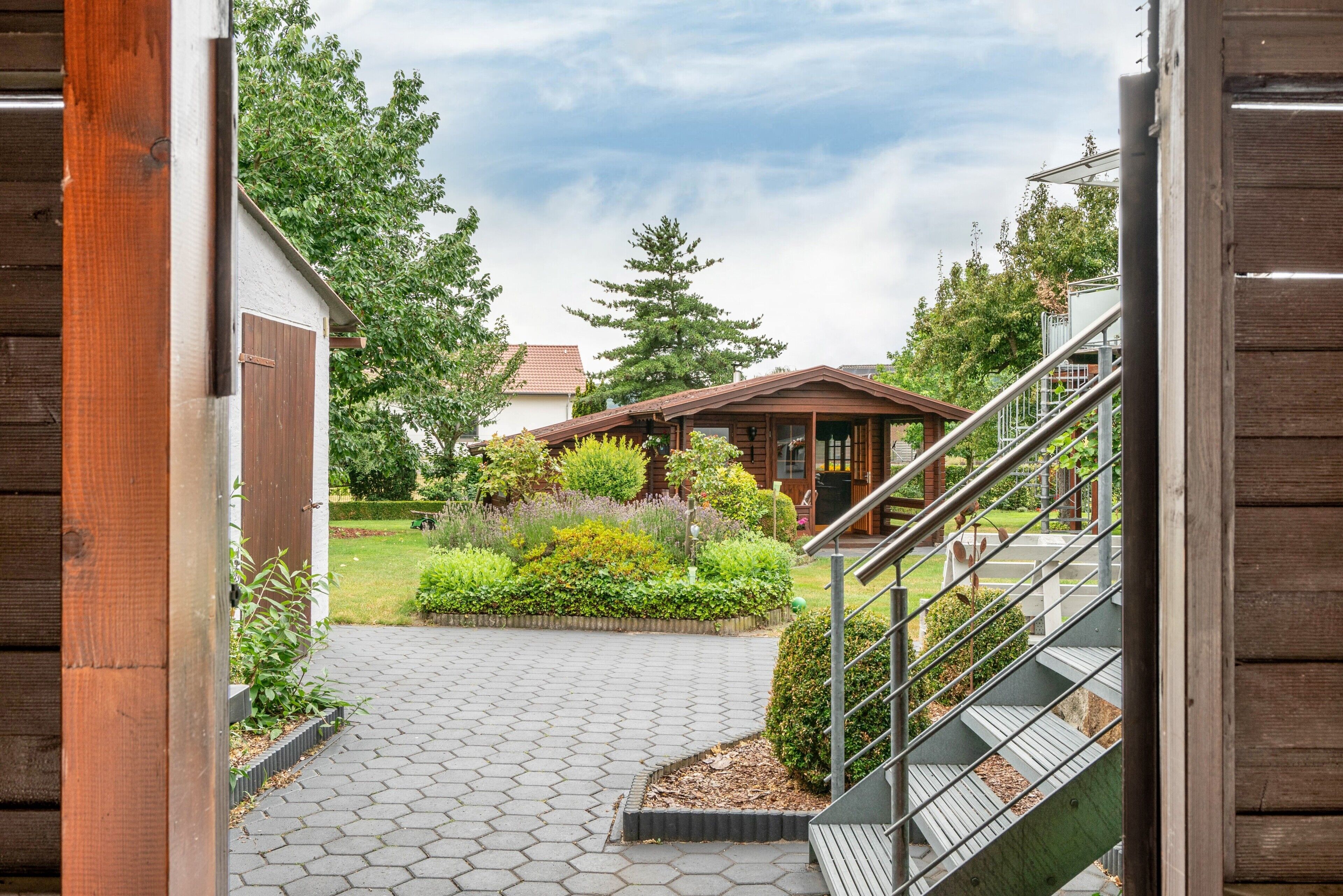 A wooden staircase leading to a covered outdoor area with a garden and a building in the background.