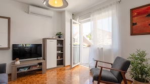 Ein Wohnzimmer mit Fernseher, Holzboden, einer Couch, einem Sessel, einem Schrank und einem großen Fenster mit Vorhängen.