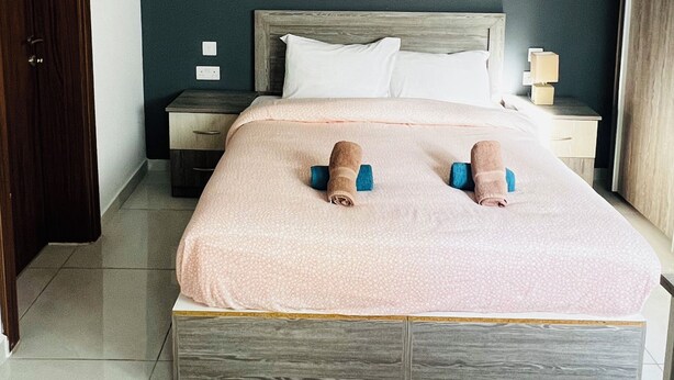 Ein ordentlich bezogenes Bett mit einer rosa Decke, einem Holzkopfteil und zwei aufgerollten Handtüchern. Ein Nachttisch mit einer Lampe und einer Steckdose ist vorhanden. Der Raum hat dunkle Wände und einen gefliesten Boden.