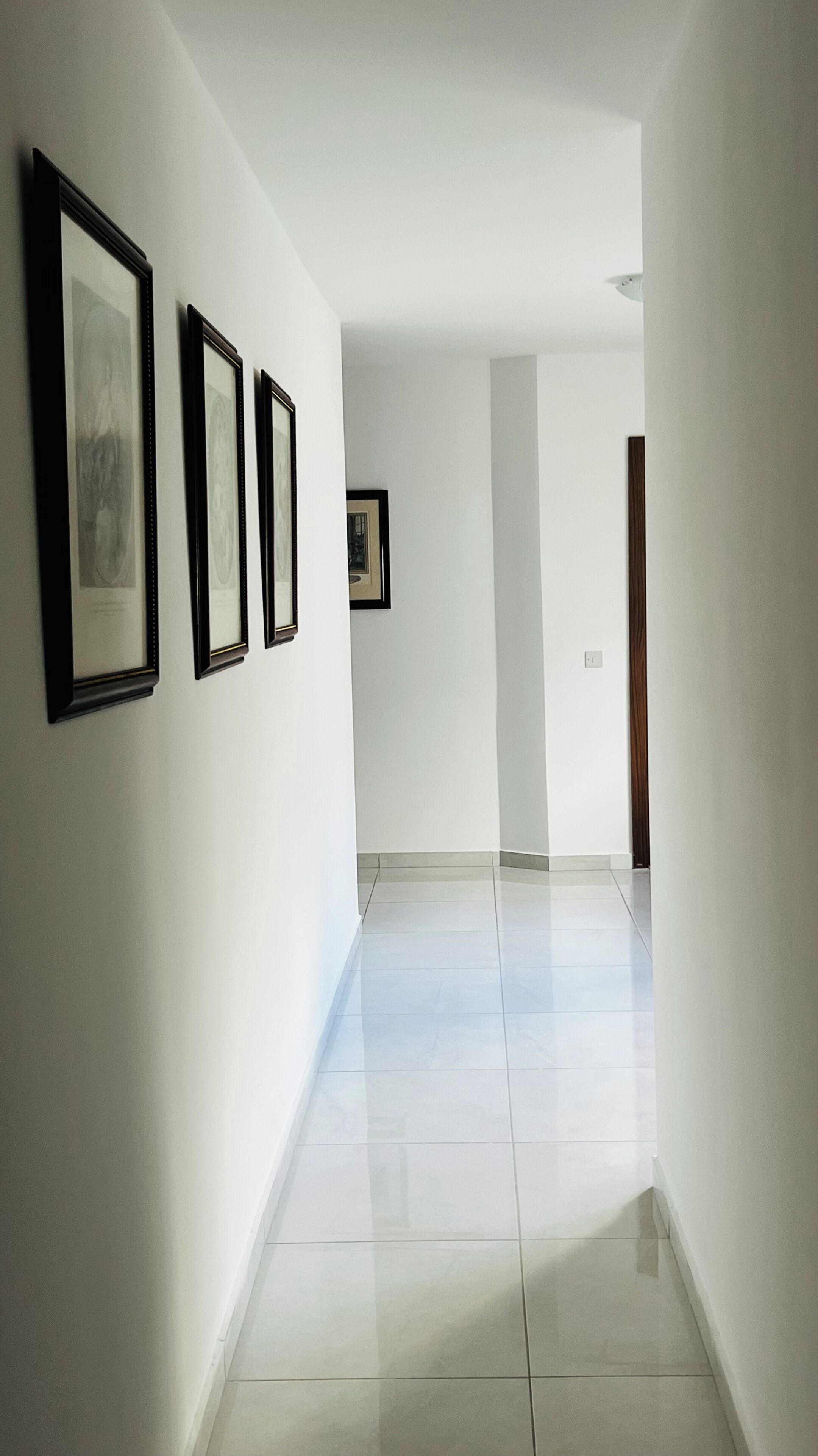 A hallway with white walls, a tiled floor, and framed pictures on the left wall.