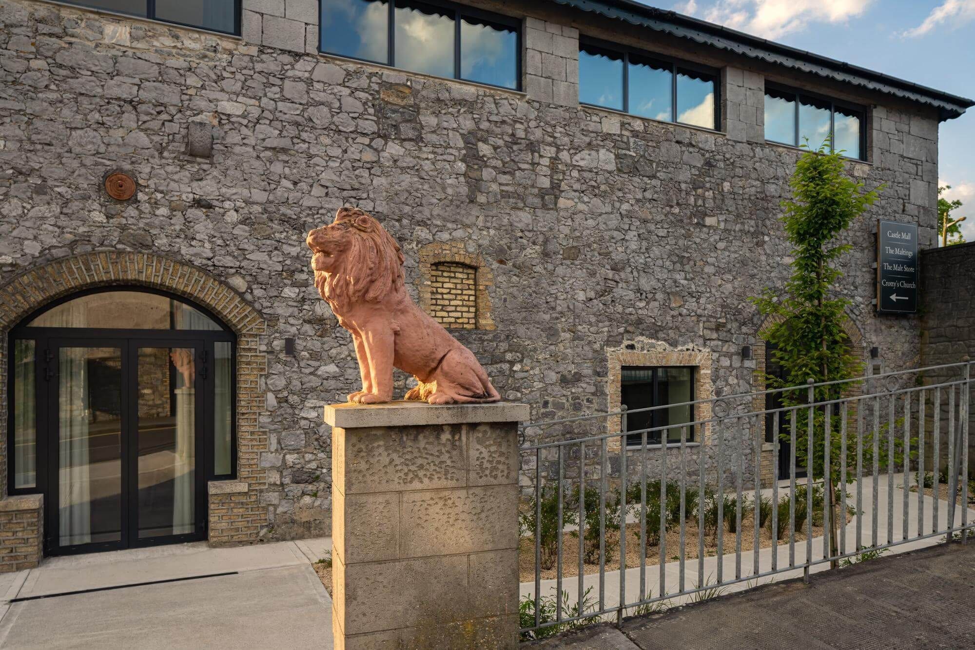 A stone building with a lion statue, a metal fence, and a sign indicating directions to Castle Mall, The Maltings, and Crotty's Church.