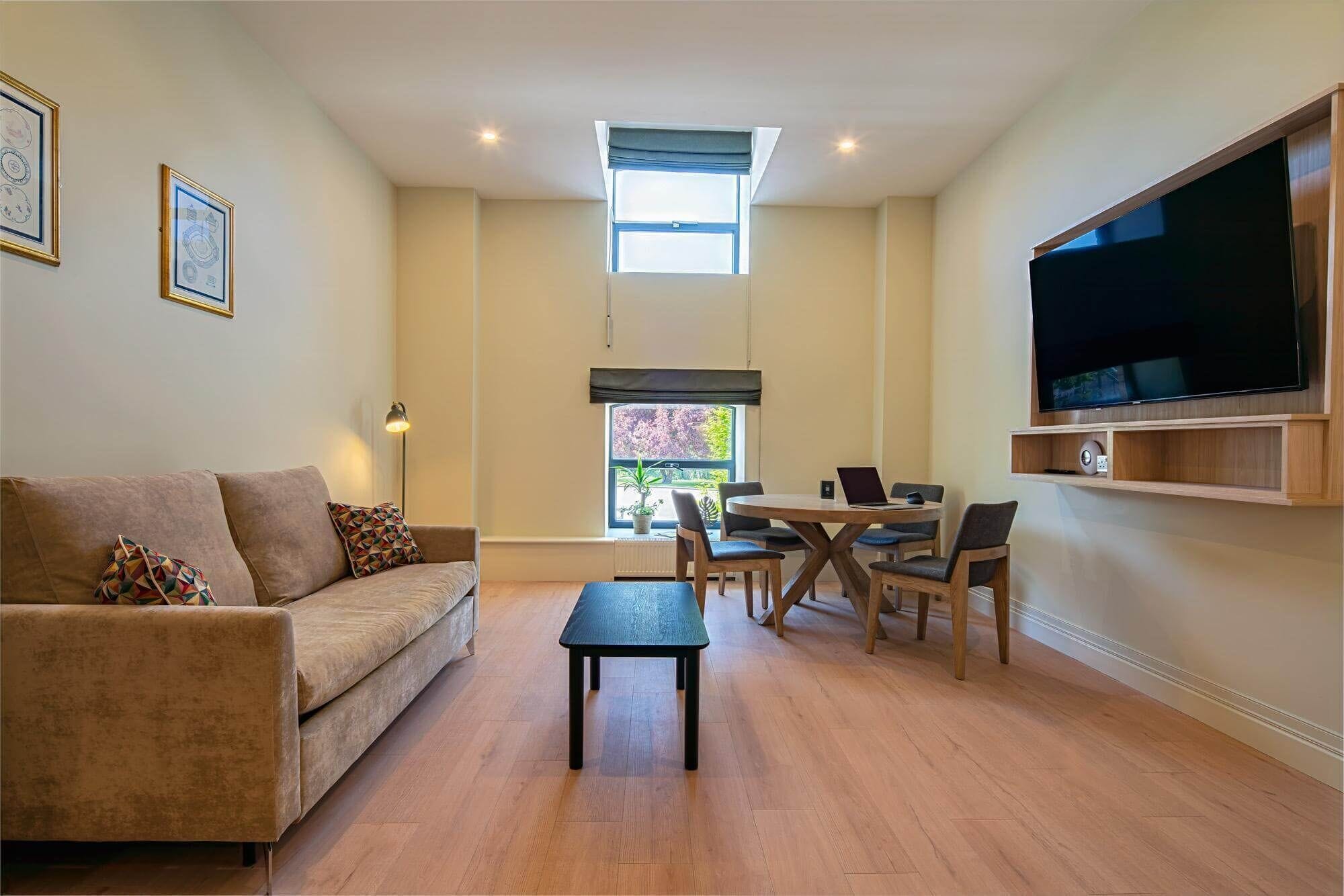 A living room with a sofa, coffee table, dining table with chairs, a TV mounted on the wall, and a window with a view of trees.