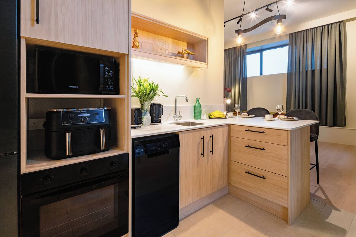 A compact kitchen with a microwave, dishwasher, wooden cabinets, and a countertop with a plant and a bottle.