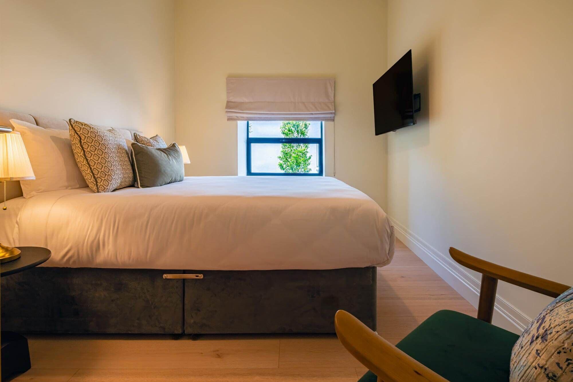 Un dormitorio con cama, mesita de noche, lámpara, ventana con persianas y un televisor montado en la pared.