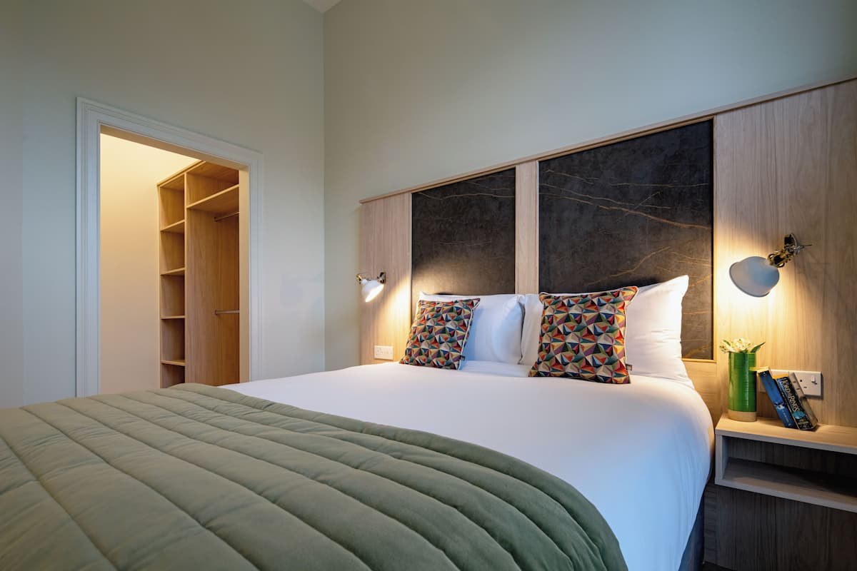 A neatly made bed with a green blanket, a wooden wardrobe, and a bedside table with a lamp and books.