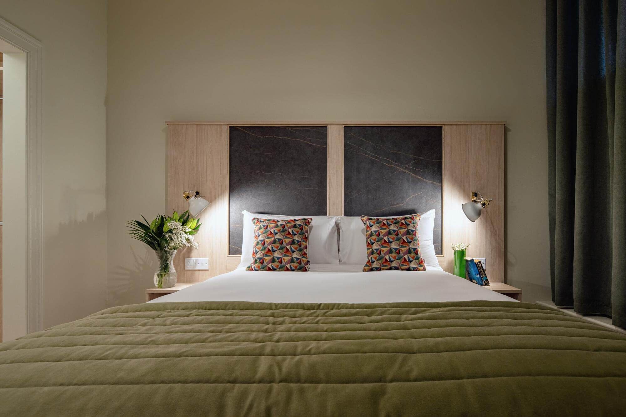 Habitación de hotel con cama, mesita de noche, un florero y una lámpara de pared.