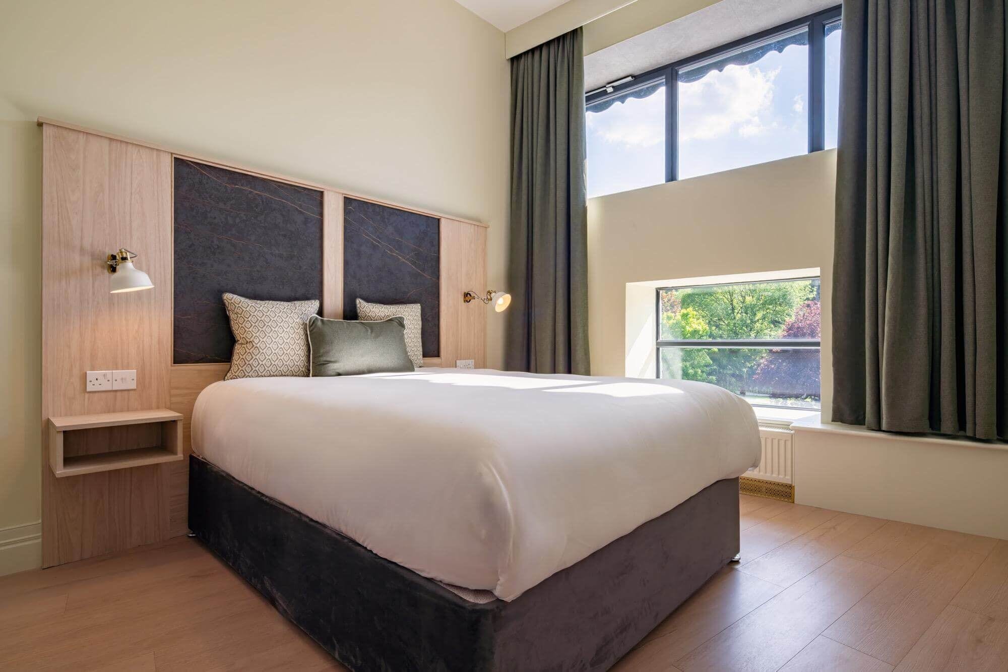 A modern bedroom with a large bed, built-in headboard, bedside lamps, and a window with curtains.
