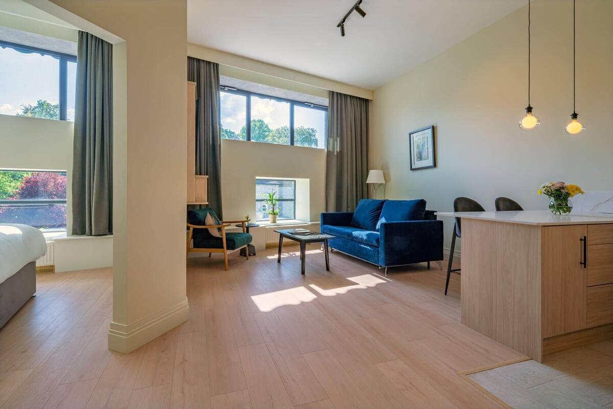 A modern hotel room with a bed, a blue sofa, a small table, a desk, and a view of greenery outside.