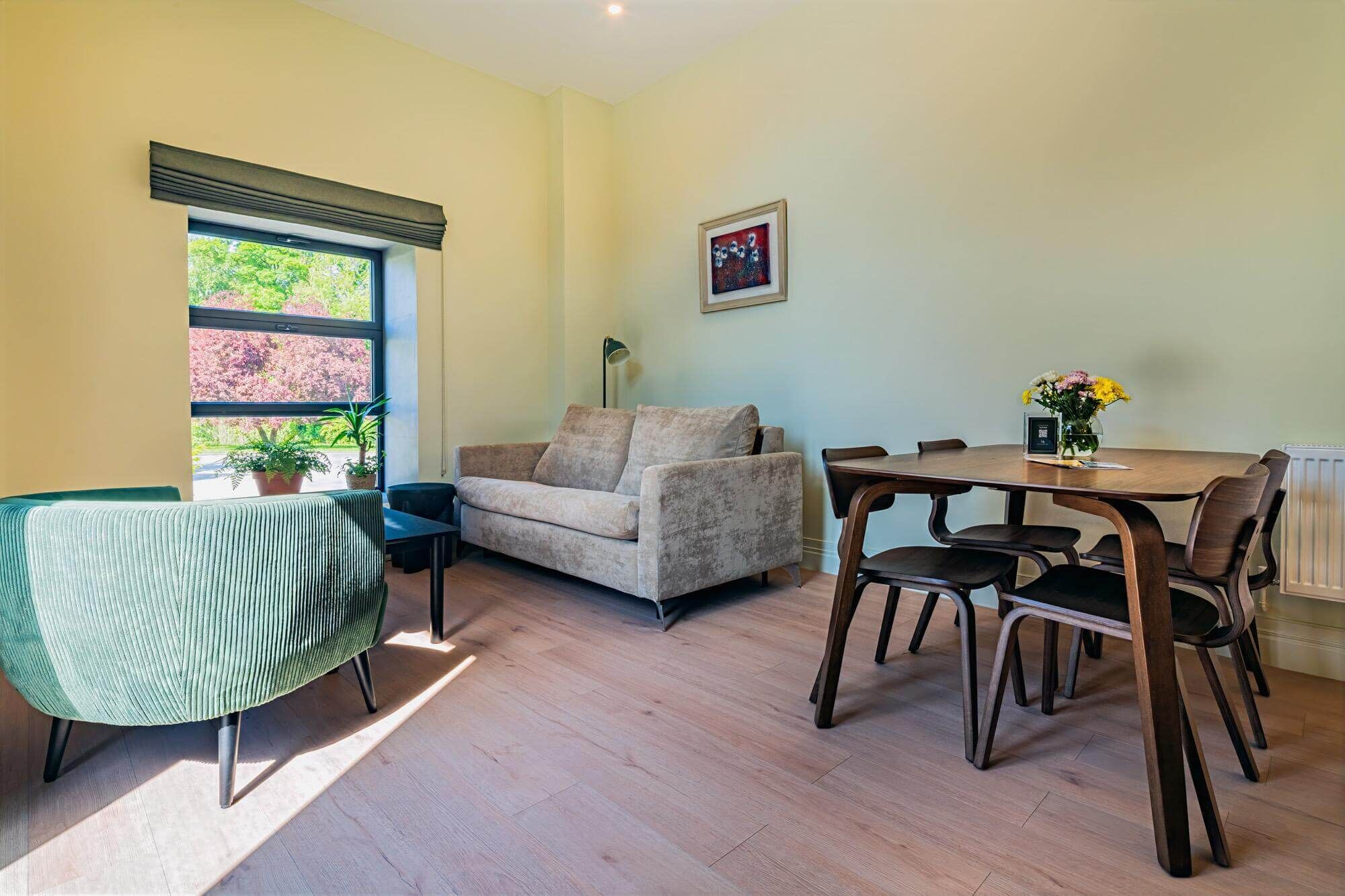 Una sala de estar con un sofá, una mesa de comedor con sillas, una silla verde, una planta en maceta y un cuadro en la pared.