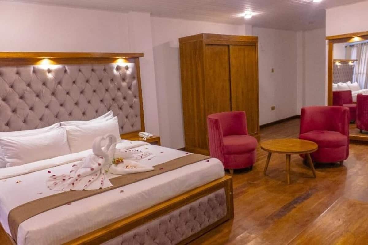 A hotel room with a large bed, a wooden wardrobe, two pink armchairs, a small round table, and a mirror.