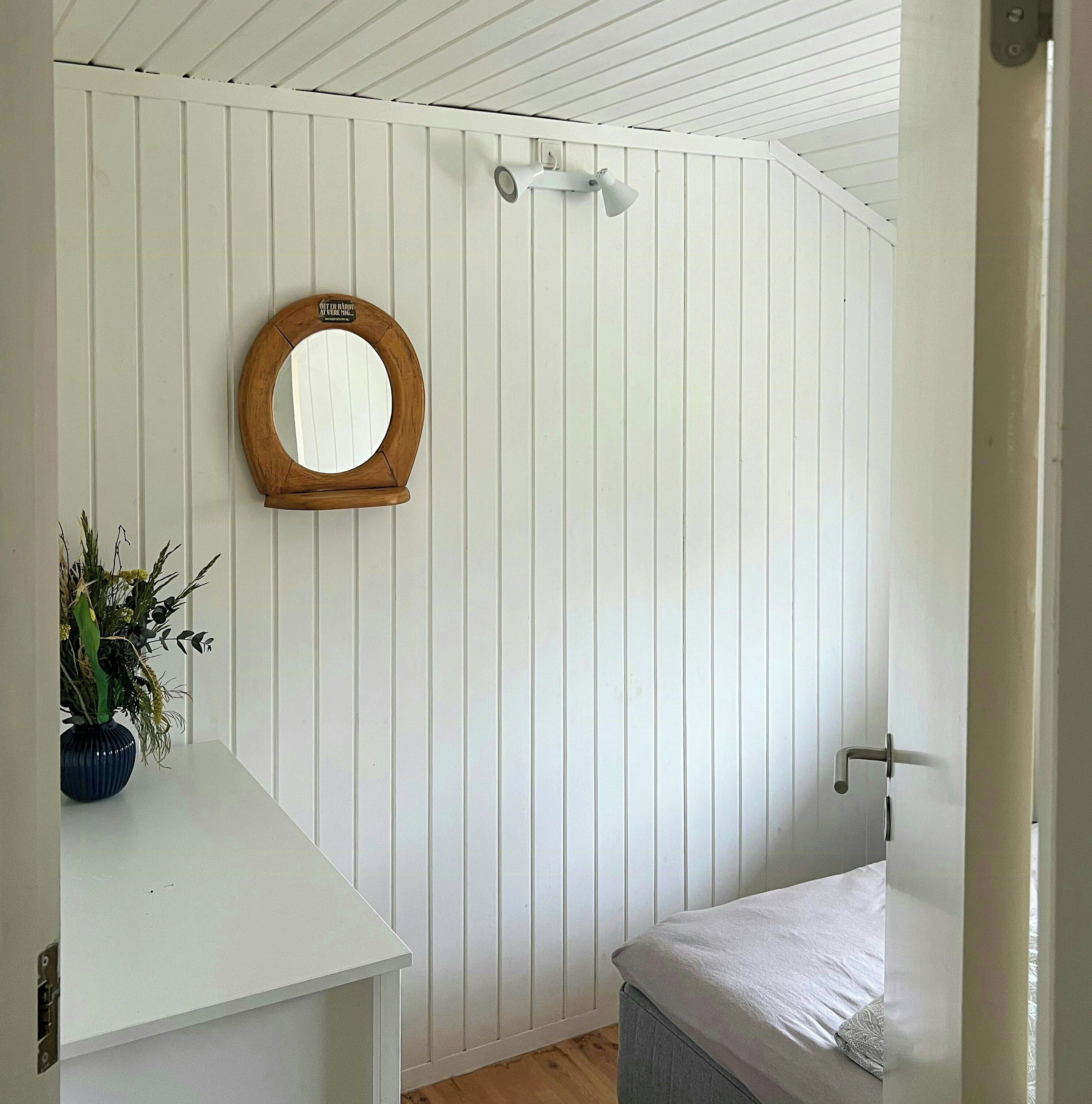 A room with a white paneled wall, a round mirror, a small table with a vase, and a bed with a white mattress and gray blanket.