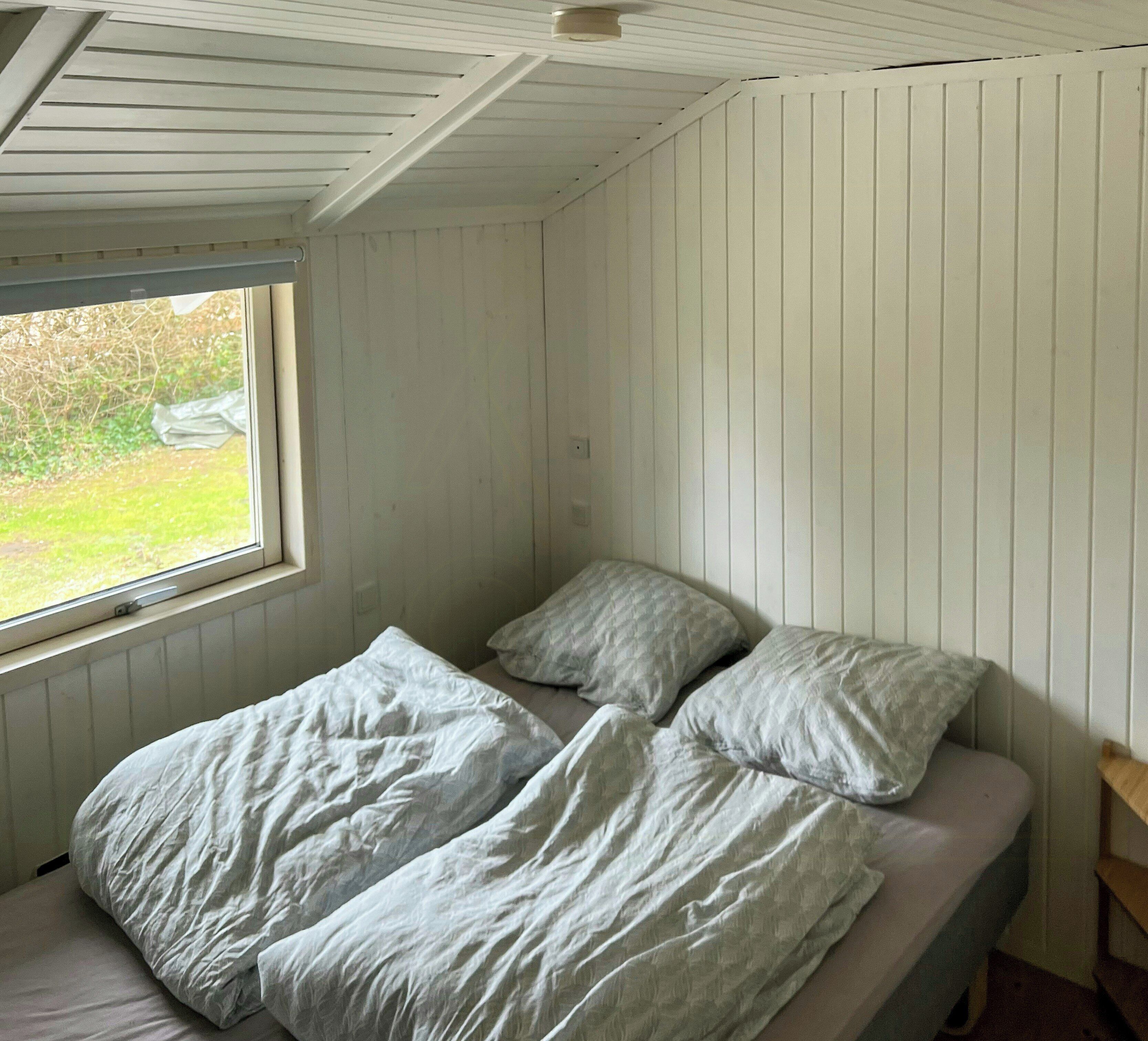 A small, single-bed room with a window, white walls, and a wooden ceiling.