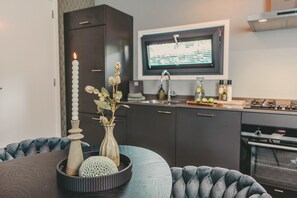 A modern kitchen with dark cabinetry, a built-in oven, and a television mounted above the sink. - KNUS Veluwe I Cozy design cottage in nature (Epe)