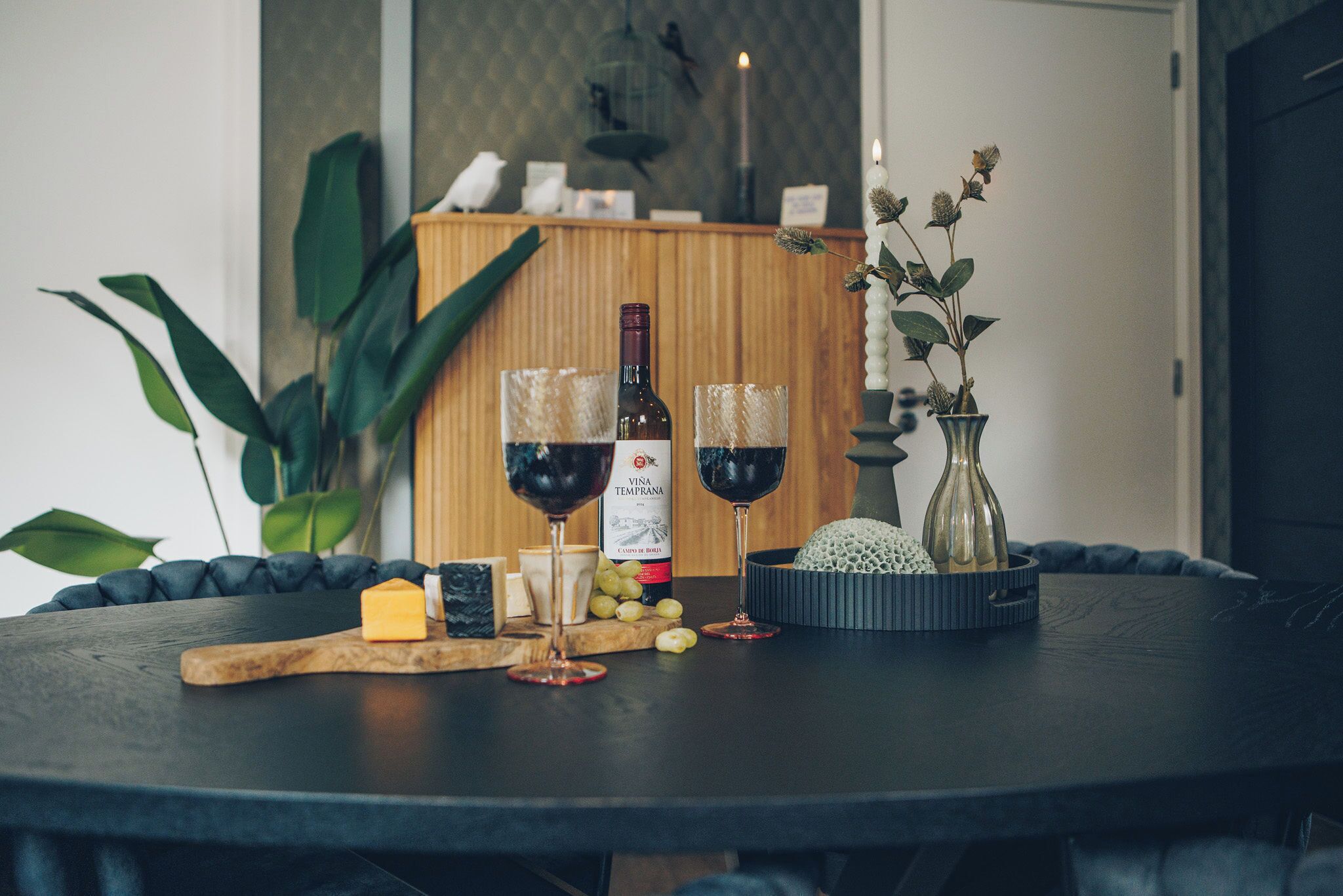 Een ronde tafel met twee glazen rode wijn, een fles wijn, kaas, druiven en een decoratieve vaas.