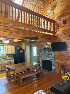 Un interior de cabaña con una mesa de comedor de madera, una chimenea de piedra, una cocina con armarios de madera y un balcón con barandillas.