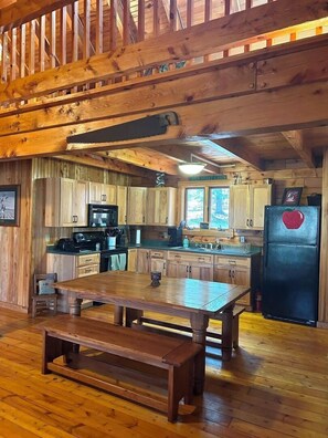 A cabin-style kitchen with wooden cabinets, a dining table, and a refrigerator. - Picture Rocks Log Cabin (Picture Rocks)
