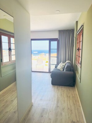 A room with a sliding glass door leading to a balcony, a grey sofa, and a wooden floor. - Tranquil Hillside Apartment – Sea View, Pool & Mountains – Los Cristianos (Arona)