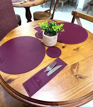 Mesa de madeira redonda com jogos americanos roxos, uma pequena planta em vaso e talheres sobre um guardanapo.