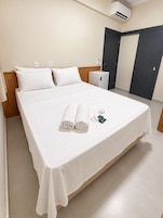 Quarto de hotel com cama, pequena escrivaninha, aparelho de ar condicionado e uma porta.
