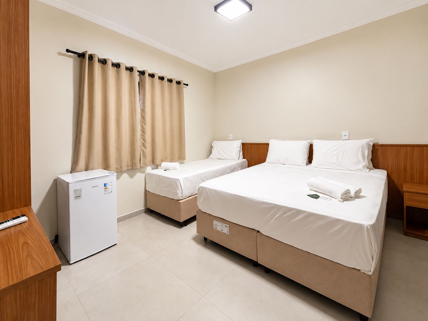 A hotel room with a bed, two bedside tables, a small refrigerator, and a window with curtains.