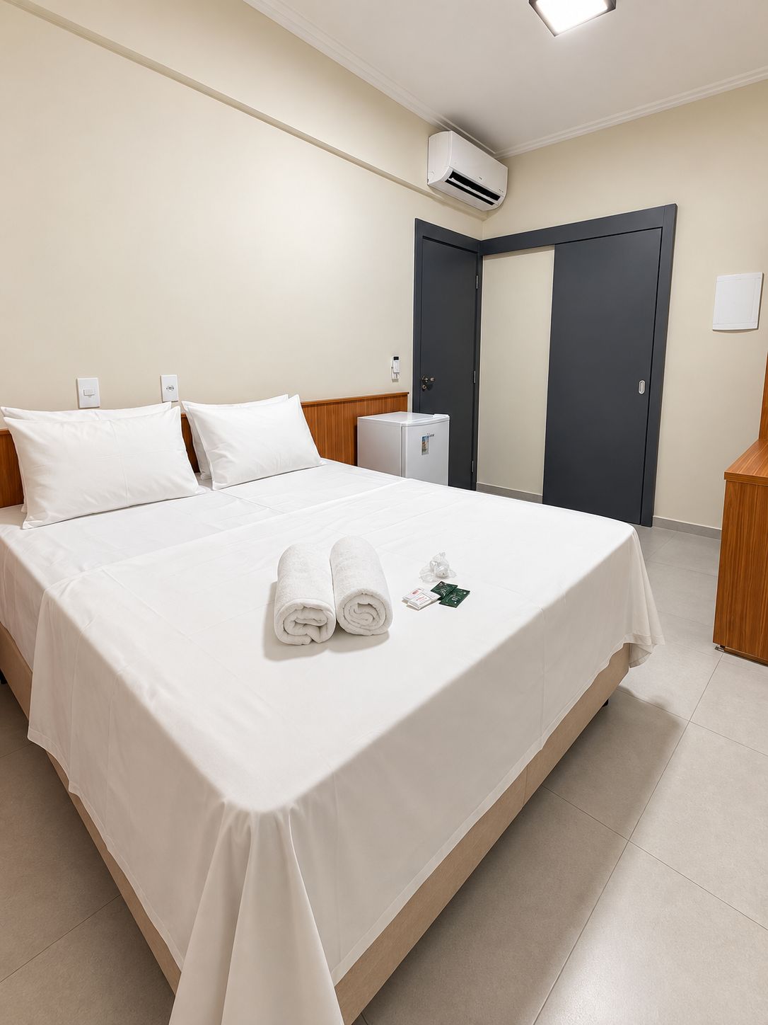 A hotel room with a bed, a small refrigerator, an air conditioning unit, and a door.