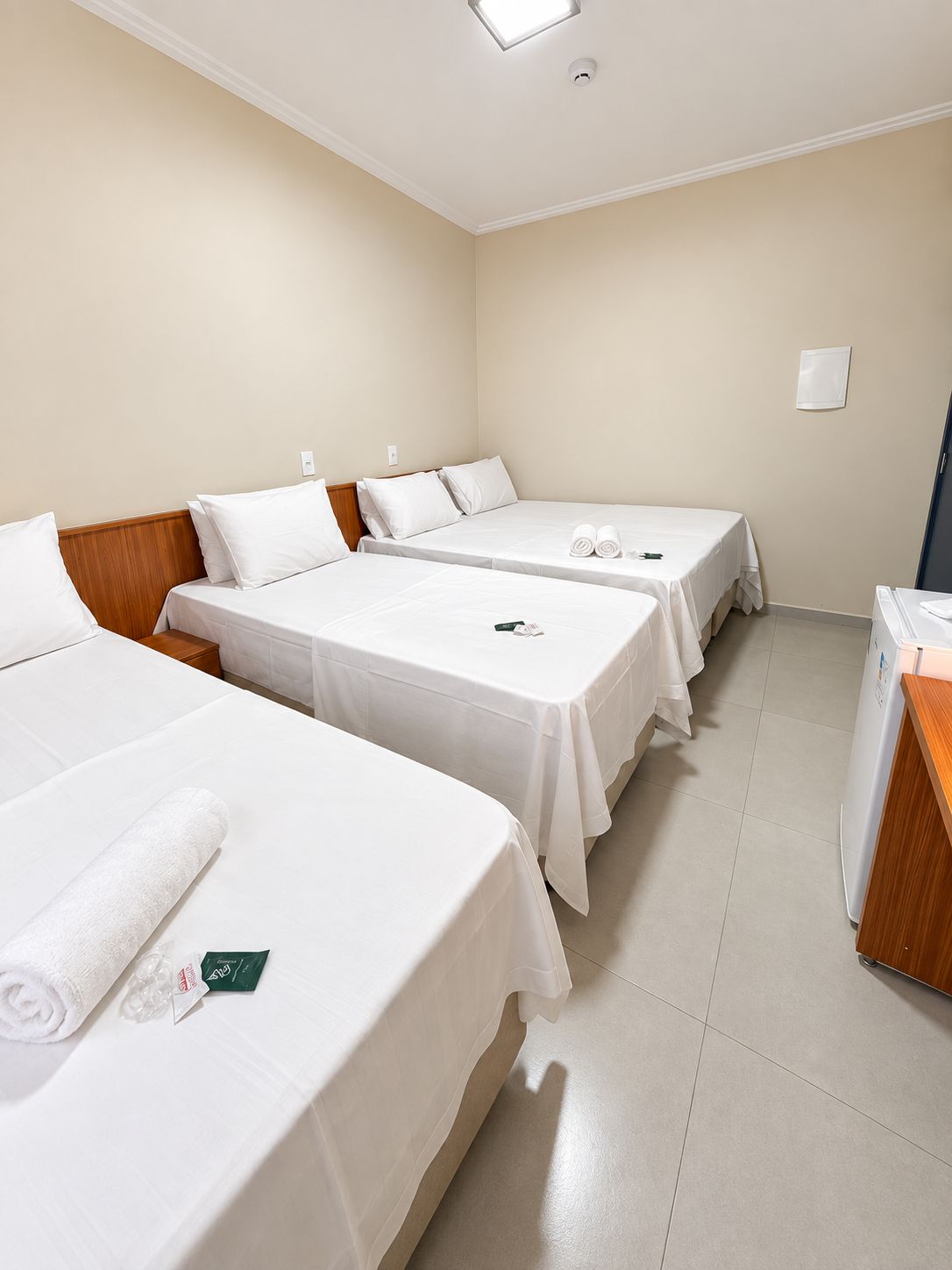 Quarto de hotel com três camas de solteiro, uma mesa de cabeceira de madeira, parede branca e luminária no teto.