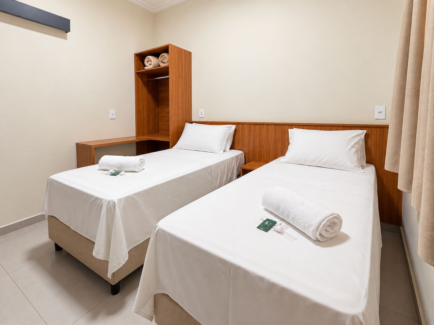 Habitación de hotel con dos camas individuales, una mesita de noche de madera y un estante con toallas.