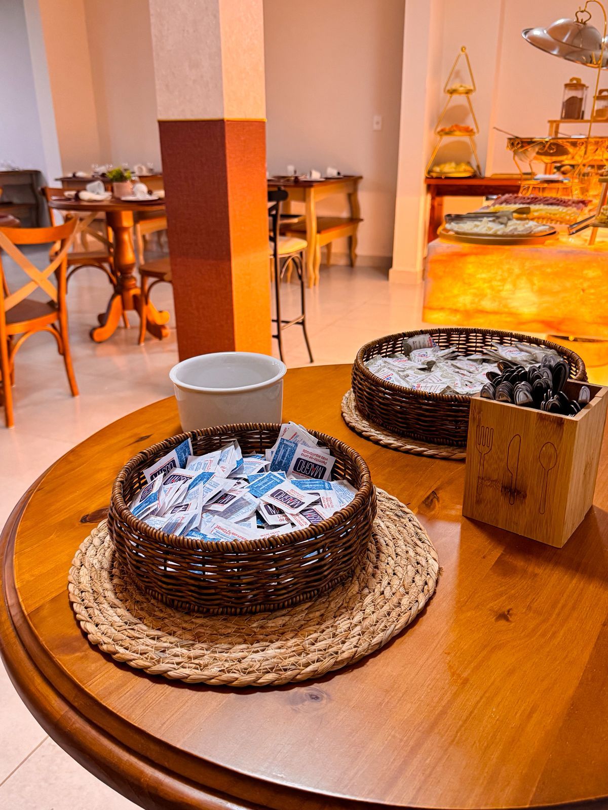 A dining area with a round wooden table, wicker baskets filled with newspapers, a white mug, and a wooden box with utensils.