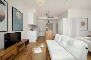 Ein modernes Wohnzimmer mit Flachbildfernseher, Couchtisch aus Holz, weißem Sofa und einer Küche im Hintergrund.