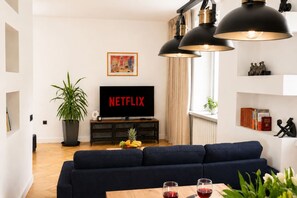 Ein Wohnzimmer mit Fernseher, einer dunklen Couch, einem Couchtisch mit Gläsern und einer Topfpflanze.