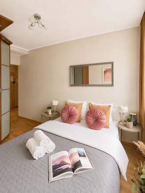 Ein ordentlich eingerichtetes Schlafzimmer mit einem Bett, Kissen, einem Nachttisch mit Lampe, einem Spiegel und einem Buch auf dem Bett.