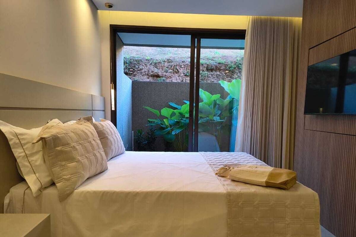Habitación de hotel con cama, televisor y puerta corrediza de vidrio que da a un balcón con plantas.