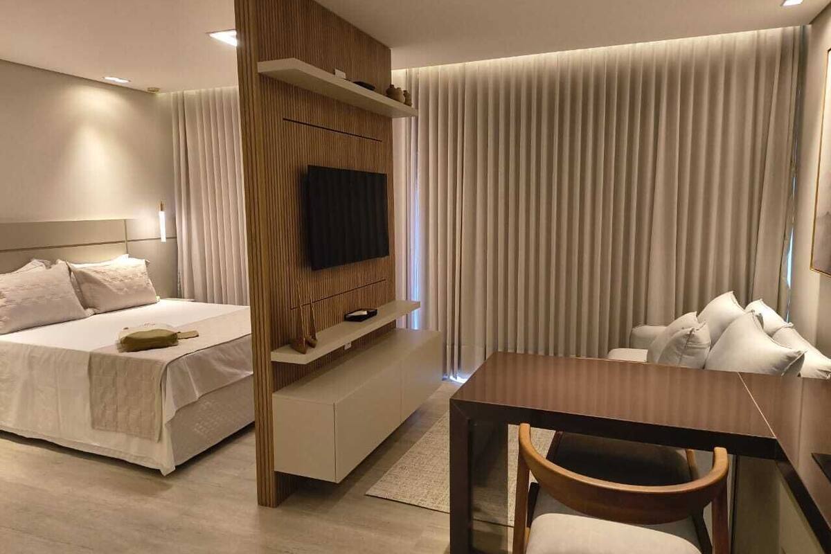 Una habitación de hotel moderna con una cama, un televisor montado en un panel de madera, una mesa de comedor con sillas y un gran ventanal con cortinas.