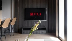 Ein modernes Wohnzimmer mit einem großen Flachbildfernseher an einer dunklen Holzwand, einem kleinen Tisch mit Stühlen und einer Vase mit Blumen.