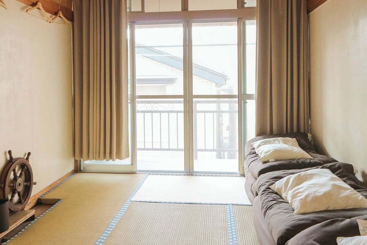 A room with tatami flooring, a bed with pillows, a wooden ship's wheel, and a sliding door leading to a balcony.