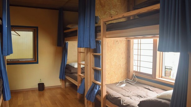 A dormitory room with bunk beds, a window with curtains, a wooden floor, and a power outlet.