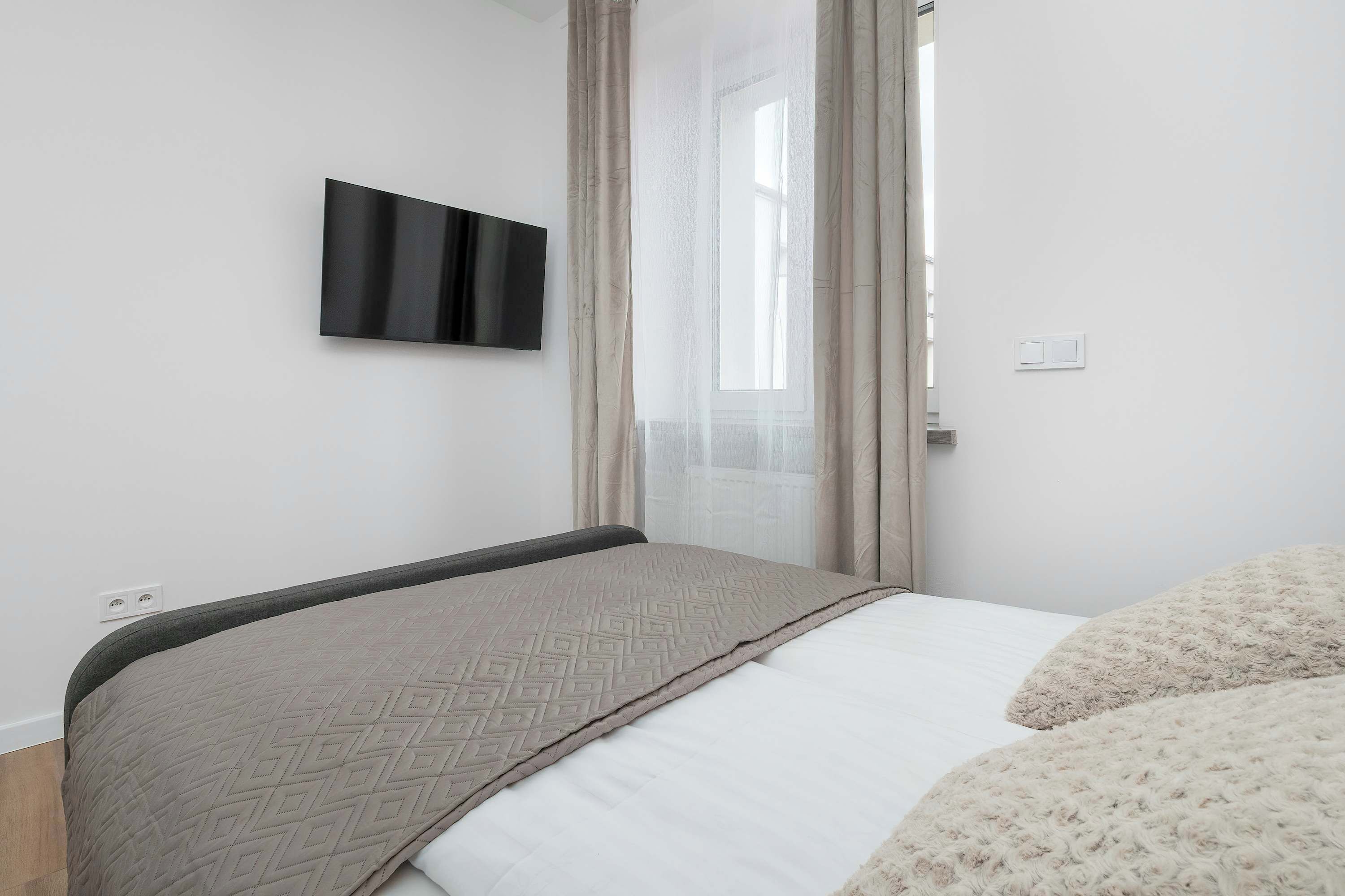 A bedroom with a bed, a TV mounted on the wall, and a window with curtains.