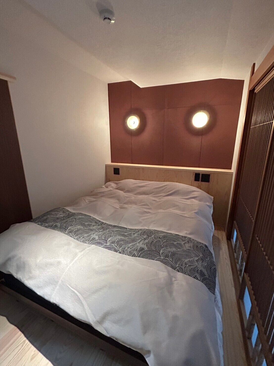 A hotel room with a bed, headboard with lights, and a wooden panel wall.